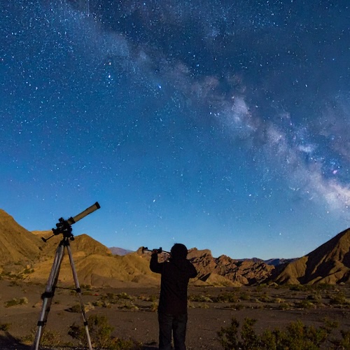 Silhouette of a young person with a telescope looking at the night sky in the dessert.