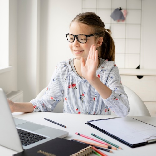 Upper elementary student wearing glasses waves and smiles during an online language arts class at a desk with books, notebook, and laptop.
