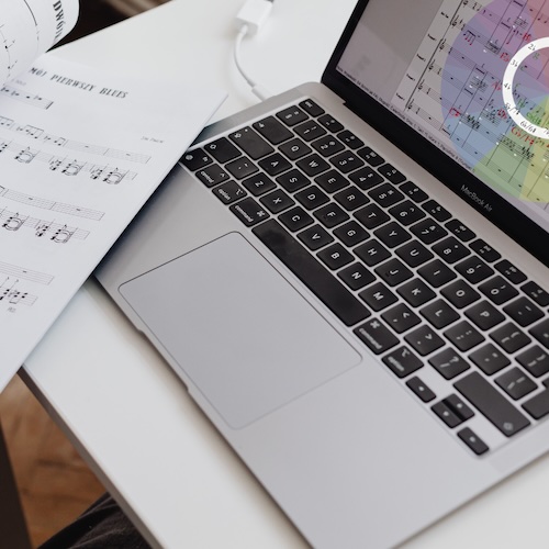 Laptop on a desk with printed sheet music and music notation, representing digital learning and creative study in Music Theory.