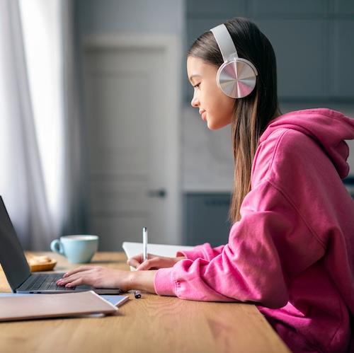 Distance education. Lady teenager in wireless headphones studying online on laptop computer at home, side view. Schoolgirl make homework assignment